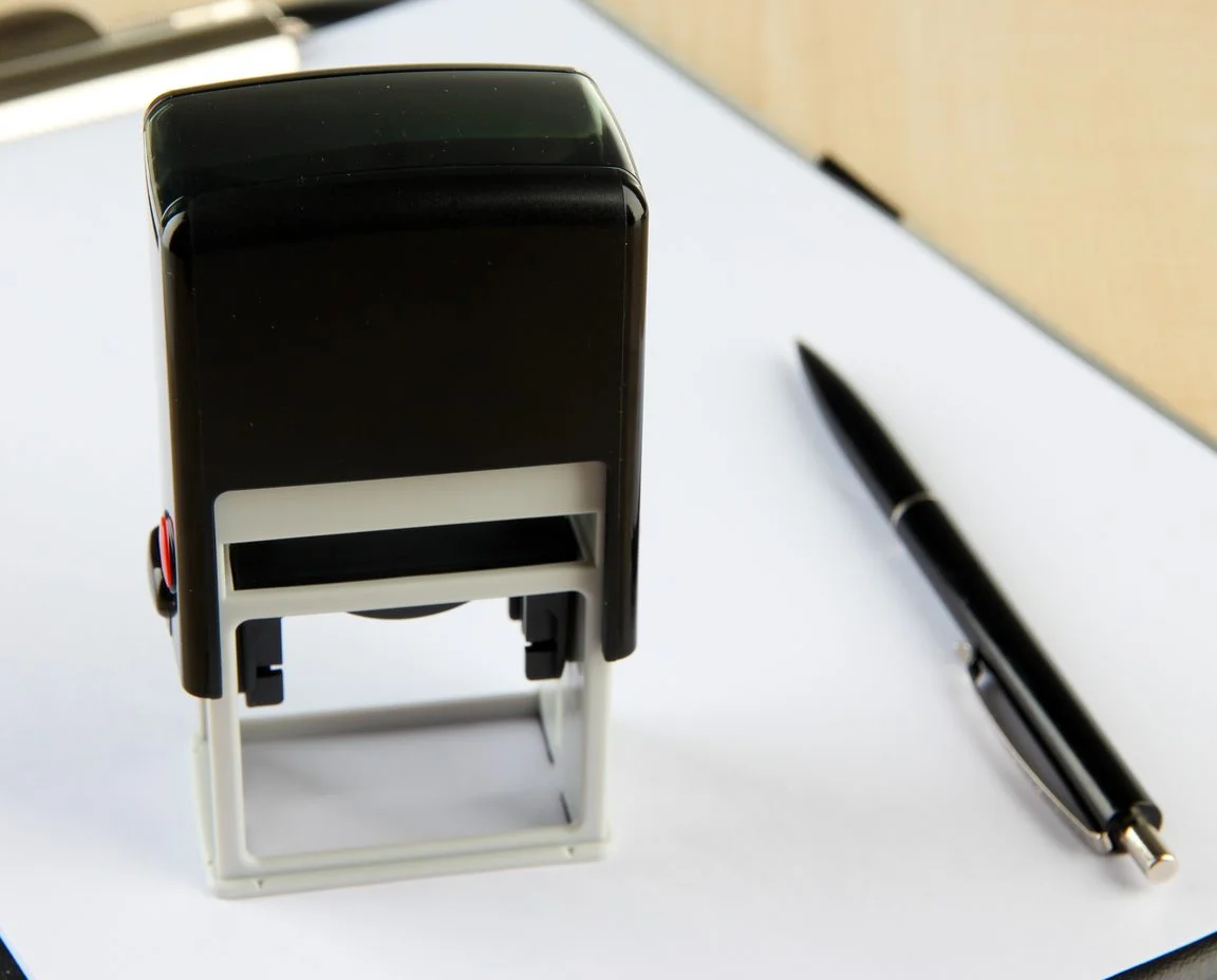Self-inking stamp and a pen resting on a blank sheet on a light wooden desk