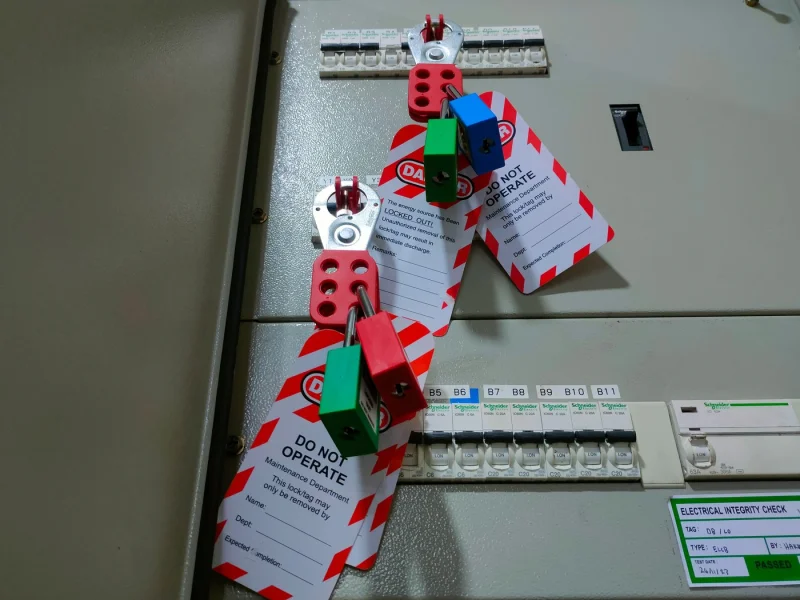 Lockout padlocks and do-not-operate tags on a switchboard, beside a dated electrical integrity check card
