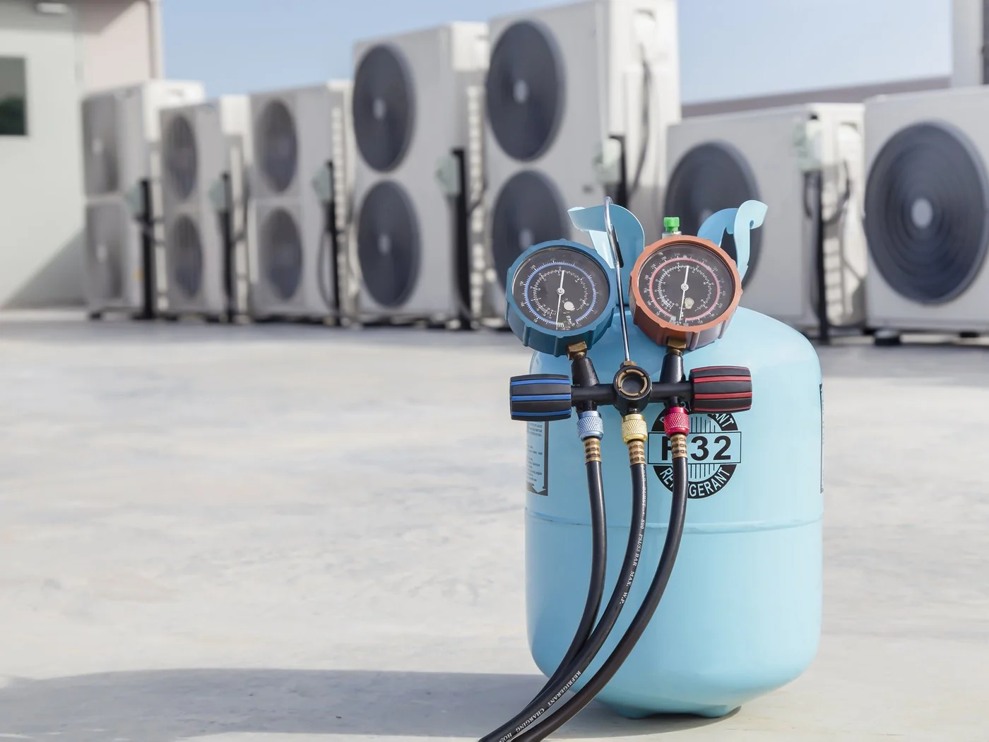 R-32 refrigerant cylinder with a manifold gauge set attached, on a rooftop in front of rows of condenser units