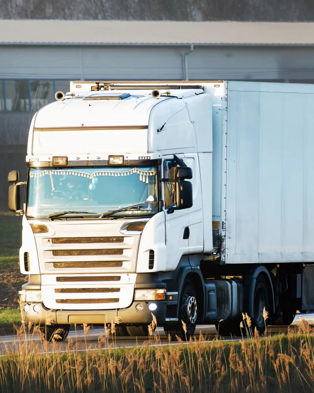 Single white tractor unit coupled to a box trailer, parked on hardstanding beside long grass